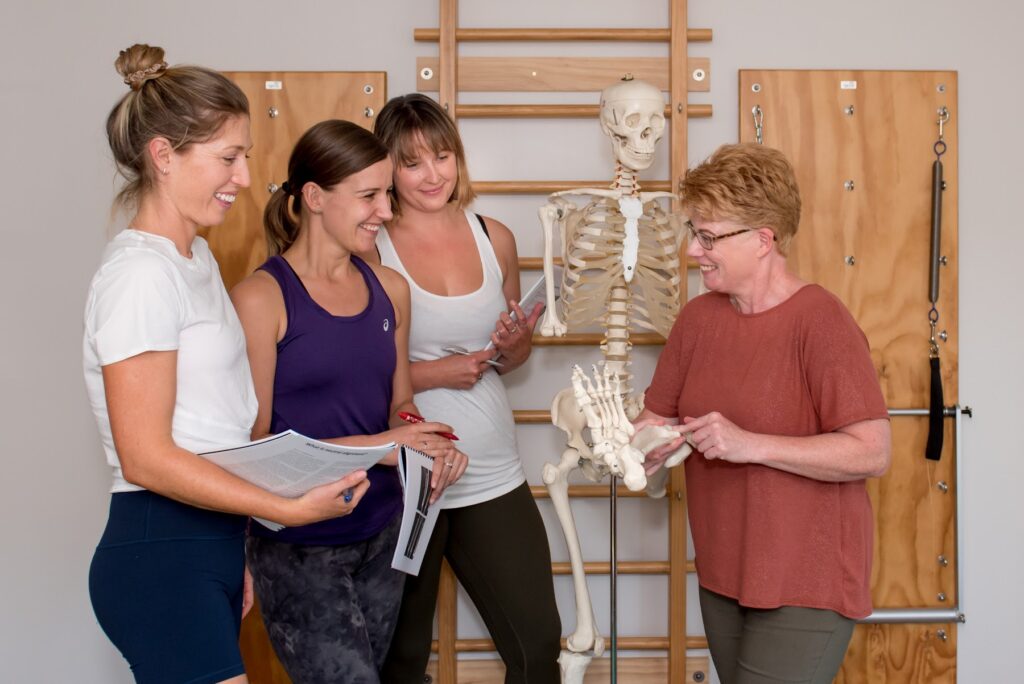 Pilates teachers studying a skeleton and anatomy charts during professional development
